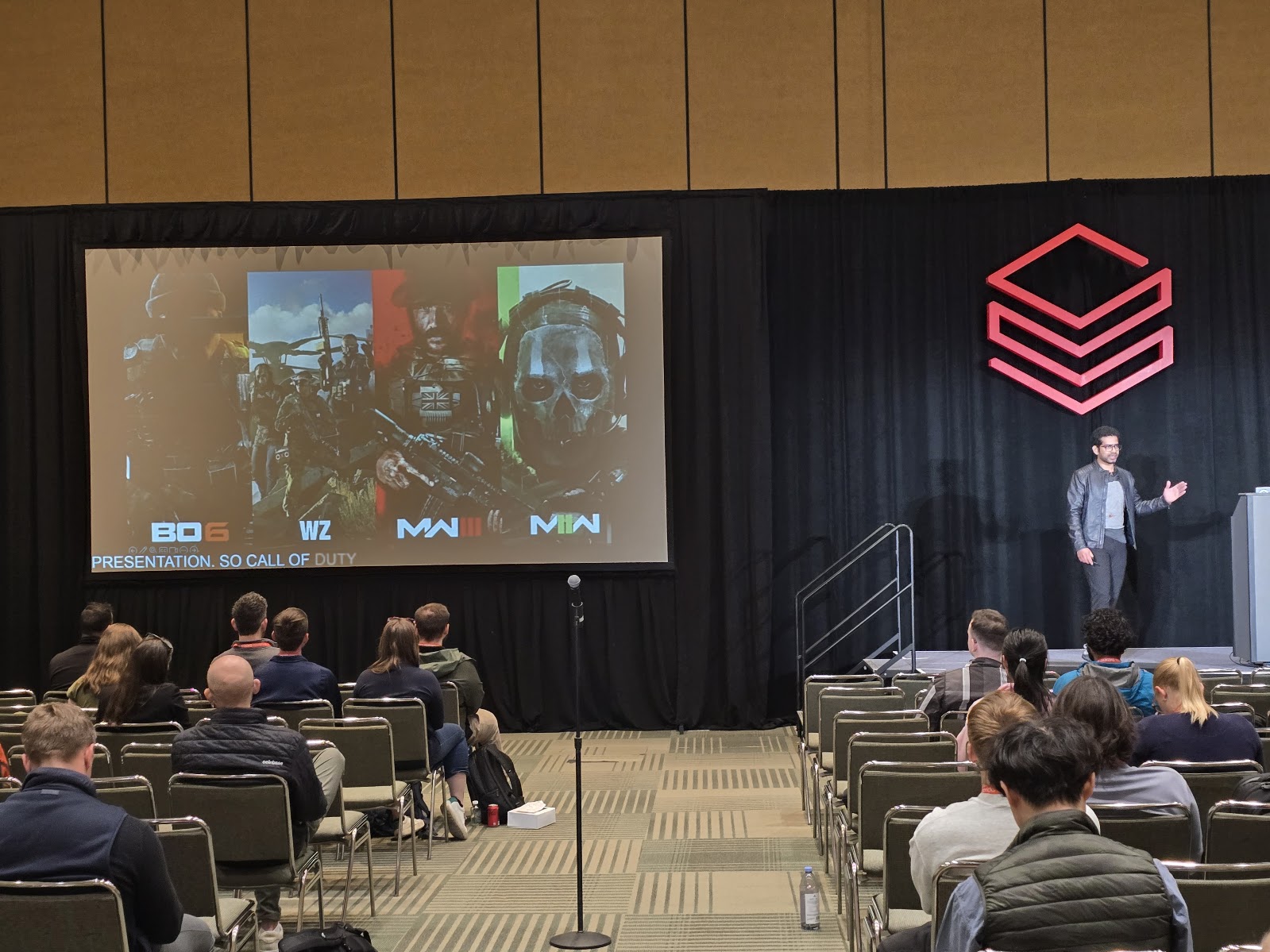 Speaker on stage beside a Databricks logo, presenting to a seated audience with Call of Duty key art on the projection screen